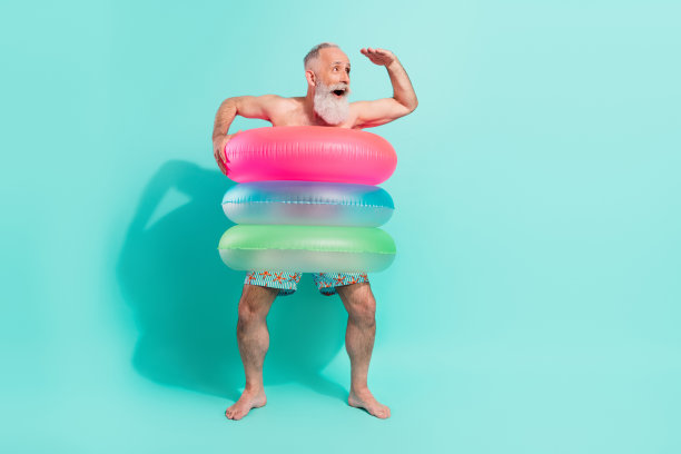 照片：一位穿着短裤、露出上身的有趣甜美退休老人，站在漂浮的圆圈内，望着远方的空白空间，背景是孤立的蓝绿色。图片下载