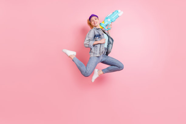 粉色背景下，戴着遮阳帽、穿着牛仔夹克的时髦金发千禧一代女士手持水枪的全身照图片下载