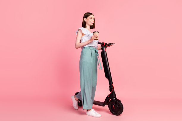 年轻女孩的全身侧面照片，快乐积极的笑容，骑着电动滑板车边走边喝咖啡，孤立在粉红色背景上图片下载