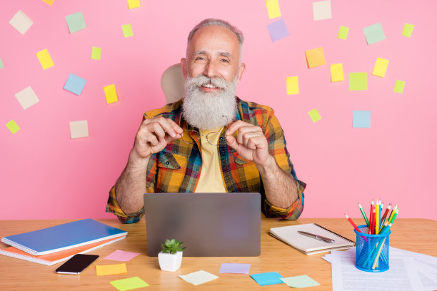 照片：一位穿着格子衬衫、认真专注的可爱老先生，坐在桌旁，用现代小工具在粉色背景上打字，背景有贴纸。图片下载