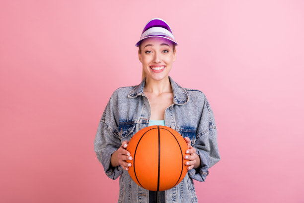 照片：一位金发千禧一代女士，穿着牛仔夹克和遮阳帽，打着篮球，在粉色背景前，表情惊讶图片下载