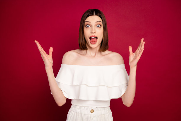 照片：一位愤怒的棕色头发的千禧一代女士，双手举起，穿着白色露肩上衣，喊叫，孤立在红色背景上图片下载