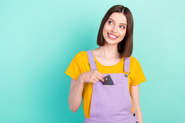 一张年轻漂亮的、充满梦想的、快乐的女性照片，她茫然地看着远方，一只手拿着信用卡放在口袋里，背景是孤立的青色。图片下载