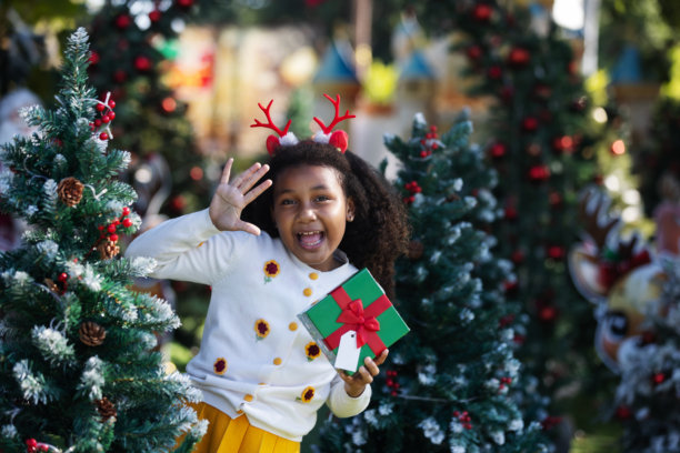 一个快乐的孩子戴着驯鹿角，开心地挥手，手里拿着圣诞礼物，周围是装饰过的松树。这张照片捕捉到了节日气氛、假日兴奋、童年快乐和户外季节性庆祝的景象。图片下载