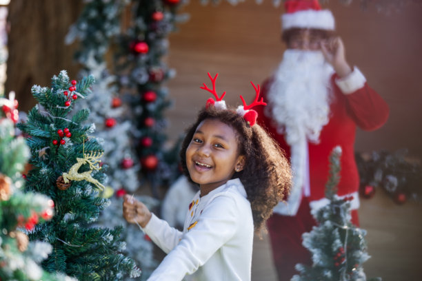 一个戴着驯鹿角的快乐孩子在户外装饰圣诞树，背景中有圣诞老人。这张图片捕捉了节日的喜悦、假期的魔力、家庭传统、童年幸福和季节性庆祝活动。图片下载
