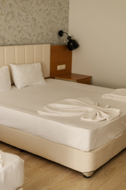 一张铺着白色床单的大床的酒店房间。床上放着两条毛巾。床边配有床头柜和台灯。装饰简约而整洁。图片下载