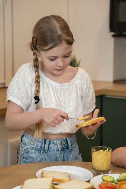 女孩正在厨房台面上仔细地将黄色芥末酱涂抹在白面包片上，准备制作三明治，旁边摆放着蔬菜和火腿等其他配料。竖幅照片图片下载