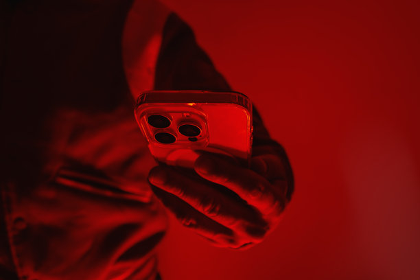 男人的手中拿着一部现代智能手机，手机在红色工作室灯光下。该图片概念涉及社交沟通、数字网络安全、黑客活动和暗网。图片下载