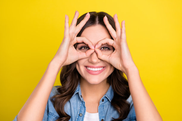 身穿牛仔衬衫、戴着眼镜、面带微笑的年轻女性，双手举起，在黄色背景前嬉戏玩耍的特写照片图片下载