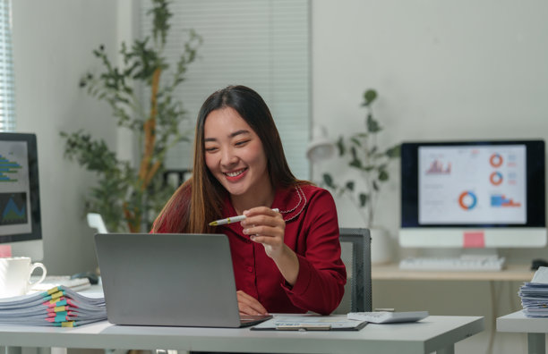 一位年轻的亚洲女商人正在办公室的办公桌前，用笔记本电脑参加视频会议，她微笑着，手里拿着一支笔指向屏幕。图片下载
