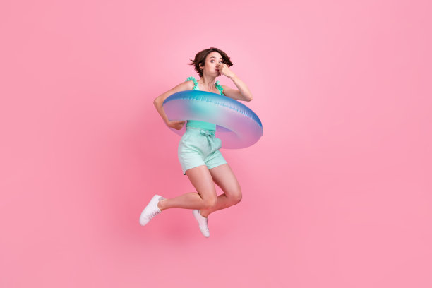 身穿蓝绿色上衣和短裤的年轻女子，留着清爽短发，身旁有游泳圈，在粉色背景前全身跳跃的照片图片下载