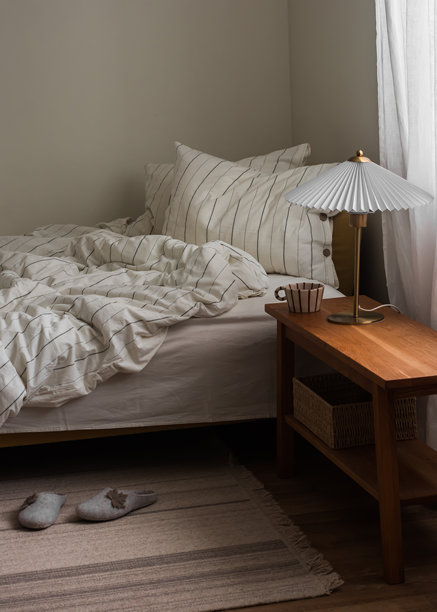 舒适的卧室 - 一张未整理的床、一盏台灯、一杯咖啡、地毯、拖鞋和一张木制床头柜图片下载