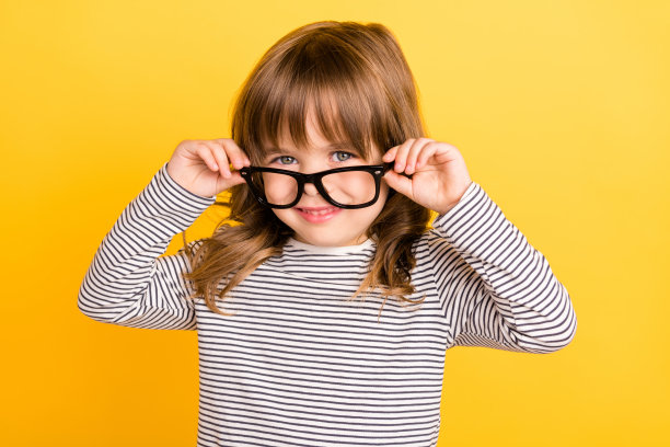 照片：一位戴着条纹衬衫、金色短发的可爱小女孩，触摸着眼镜，背景为黄色。图片下载