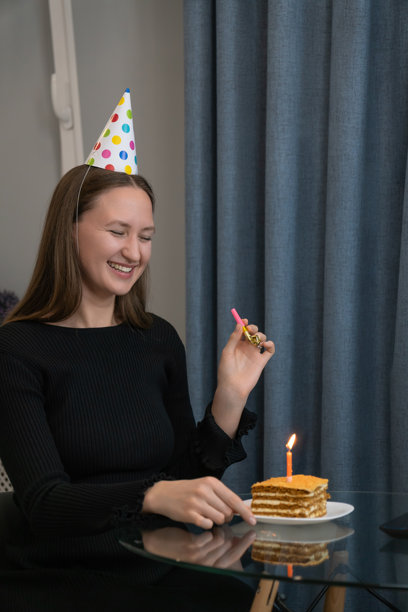 一位戴着派对帽的年轻女子正在厨房里，在生日蛋糕和点燃的蜡烛前，对着镜头吹响派对喇叭庆祝生日。竖幅照片图片下载