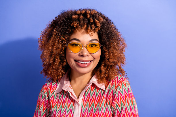 一位卷发、戴着黄色眼镜、身穿彩色衬衫的年轻时尚女性，在蓝色背景前摆姿势，用于时尚生活方式图库照片图片下载