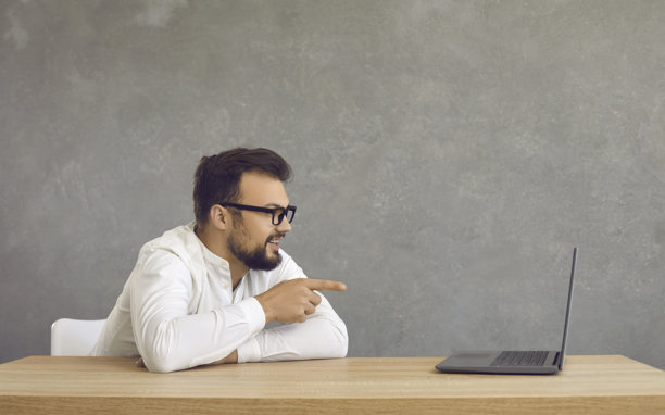 快乐的年轻男子坐在办公桌前，用手指着笔记本电脑的屏幕图片下载