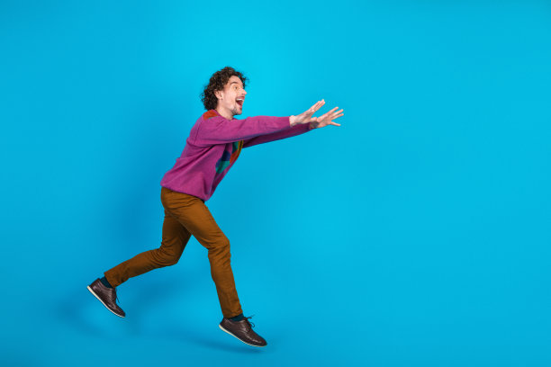 一个穿着彩色毛衣的年轻男子，脸上带着喜悦的表情，向前奔跑，背景是明亮的蓝色，适合生活方式和广告图片图片下载