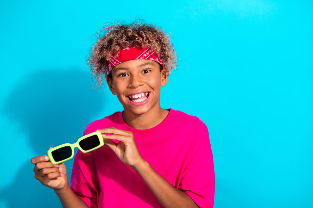 穿着粉色衬衫、戴着霓虹太阳镜的快乐小男孩对着明亮的蓝色背景微笑，呈现出有趣的时尚生活方式素材图片下载