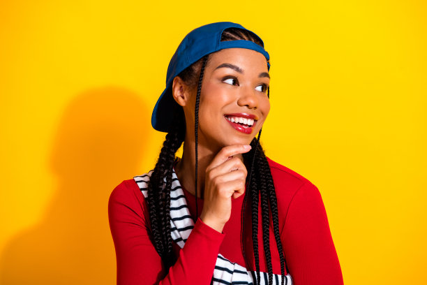 身穿红色上衣、蓝色帽子、梳着辫子的年轻时尚女性，戴着红色发带，在明亮的黄色背景前微笑图片下载