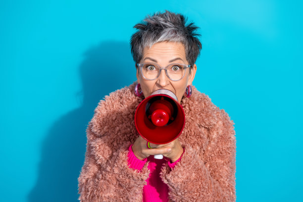 一位留着灰色短发、身穿粉色外套和毛绒夹克的年长女性，在蓝色背景前拿着扩音器讲话，画面充满活力，适合时尚和健康主题的图片。图片下载
