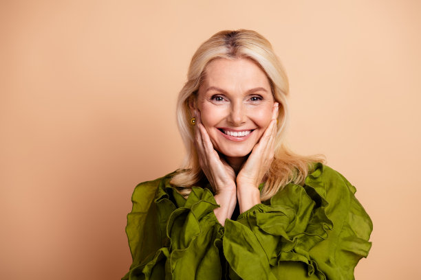 一位金发中年女性微笑着，身穿绿色荷叶边衬衫，在米色背景前摆姿势，用于健康、时尚和生活方式的素材图片图片下载