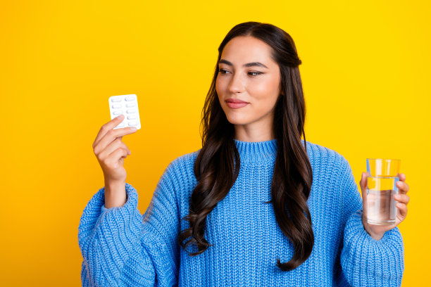 年轻的混血女子，棕色头发，身穿蓝色毛衣，手持药盒和一杯水，背景为明亮的黄色，用于健康、养生和生活方式的图库照片图片下载