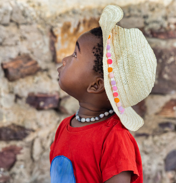 非洲村庄女孩头像，戴草帽，穿红衬衫，置身户外自然景致中，身后是石墙图片下载