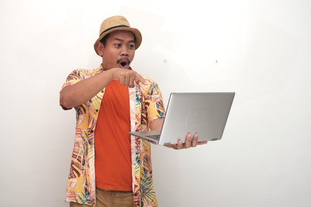 一位穿着草帽、热带风情衬衫和橙色T恤的兴奋的东南亚男性，打扮成游客，手持笔记本电脑，查看屏幕上关于促销或打折的信息，用于广告宣传。图片下载