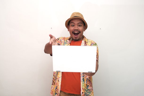 穿着草帽、色彩鲜艳的热带衬衫和橙色T恤的兴奋的东南亚男子，打扮成游客，手持笔记本电脑，查看屏幕上的促销或销售信息，用于广告宣传。图片下载