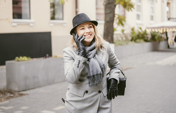 一位身穿灰色西装、戴着帽子、围巾和手套的职业女性，在商务午餐后，一边走在秋季的城市街道上，一边用智能手机通话。生活方式、通讯、商业街和城市生活概念。图片下载
