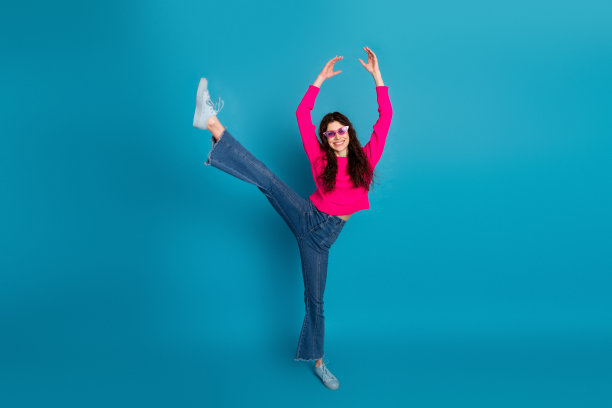 身穿粉色针织衫的年轻女子在蓝色背景前起舞踢腿，展现休闲时尚与活力生活方式，适用于现代广告和创意素材图图片下载