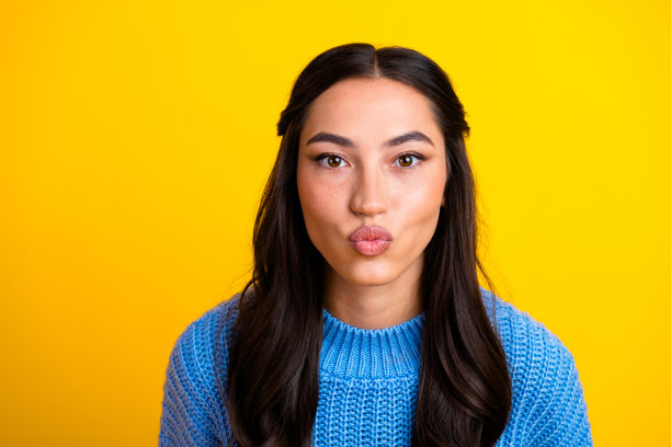 年轻的混血女子穿着蓝色毛衣，在明亮的黄色背景前噘嘴，作为休闲生活时尚模特拍摄的素材照片图片下载