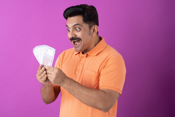 身穿橙色T恤的印度中年男子展示印度100卢比纸币，背景为紫色，为库存照片图片下载