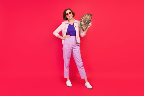 照片：一位戴着太阳镜，穿着粉色夹克的快乐退休女士，在红色背景前用扇子给自己扇风。图片下载