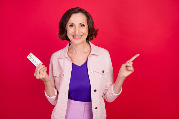 照片：一位聪慧、积极的老妇人，戴着粉色夹克，手指着药物，指向空白处，背景为红色。图片下载