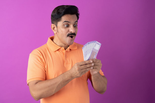身穿橙色T恤的印度中年男子展示印度100卢比纸币，背景为紫色，隔离素材图片下载