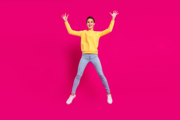 照片：一位穿着黄色套头衫的漂亮幸运年轻女子，面带笑容，双手握拳向上举起，在粉色背景前跳跃图片下载