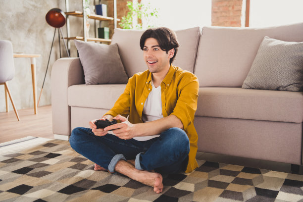 照片：一位穿着黄色衬衫的友好男士坐在舒适的沙发上，在家中室内手持电话进行视频通话并挥手。图片下载