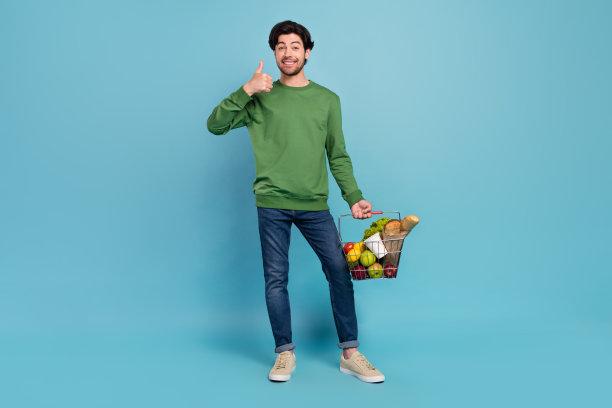年轻男子全尺寸照片，面带微笑，正在购物买菜，竖起大拇指，在蓝色背景上孤立。图片下载