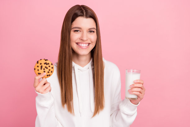 照片：一位棕色头发的千禧一代女士，戴着连帽衫，手里拿着饼干和牛奶，背景是粉色的。图片下载