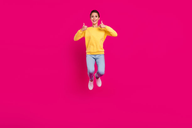 照片：一位穿着黄色套头衫的漂亮幸运年轻女子，微笑着，双手握拳向上跳跃，背景是粉红色的。图片下载