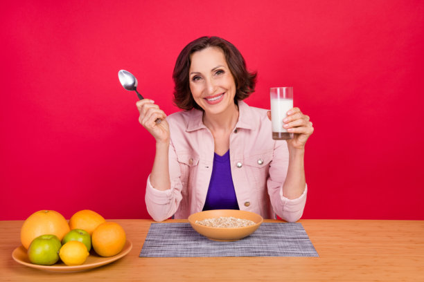 照片：一位老妇人坐在桌前，手持一杯美味新鲜的牛奶，用勺子品尝，露出灿烂的笑容，身穿粉色夹克，背景为孤立的红色。图片下载