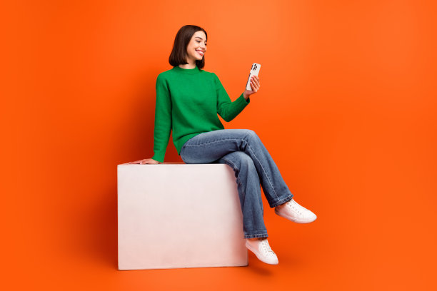 一位穿着绿色毛衣的年轻时尚女性坐在白色盒子上，背景为橙色，使用手机，这是一张明亮时尚的生活方式图片图片下载