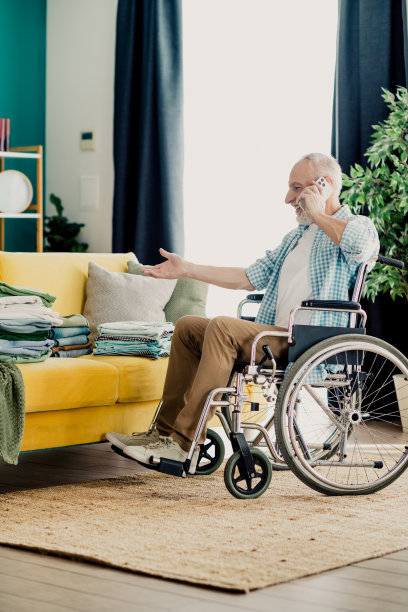 一位坐着轮椅的老人在阳光明媚的客厅里，正在打电话，传达着家庭、包容和居家日常的坚韧。图片下载