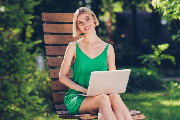照片：穿着绿色衣服、提着手袋的可爱闪亮年轻女子在城市街道外一边走一边用现代设备打字，面带微笑。图片下载