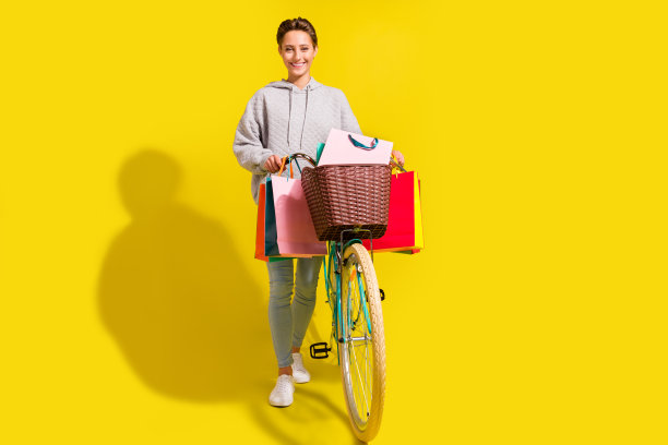 一位棕色头发的年轻女士骑着自行车，身穿毛衣、牛仔裤和运动鞋，挎着包，背景为黄色，全尺寸照片图片下载
