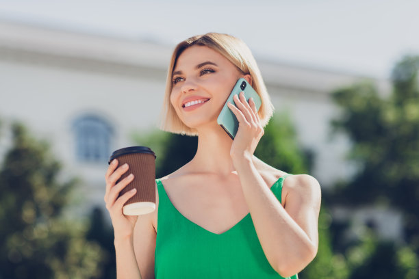 照片：穿着绿色衣服、手拿手提包的可爱闪亮年轻女子，在城市街道上行走，一边微笑一边用现代设备打字图片下载