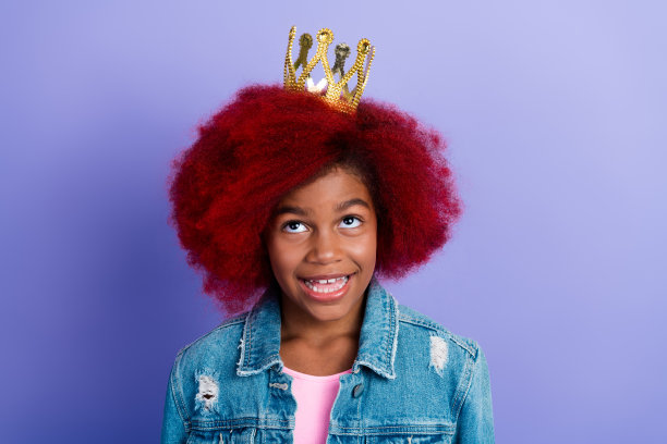 一个染着红发的年轻女孩戴着皇冠，穿着牛仔夹克，在紫色的背景前微笑，这是一张有趣的时尚生活方式的素材照片图片下载