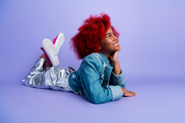 一位拥有鲜红色头发的年轻女孩，身穿牛仔夹克，在紫色的影棚背景前摆出舒适的姿势，她面带愉悦的表情，展现出时尚、趣味和复古风格。图片下载