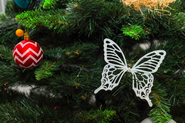 一个优雅的白色蝴蝶装饰，拥有镂空翅膀和一个带有锯齿图案的红色球体，装饰着圣诞树蓬松的树枝。图片下载